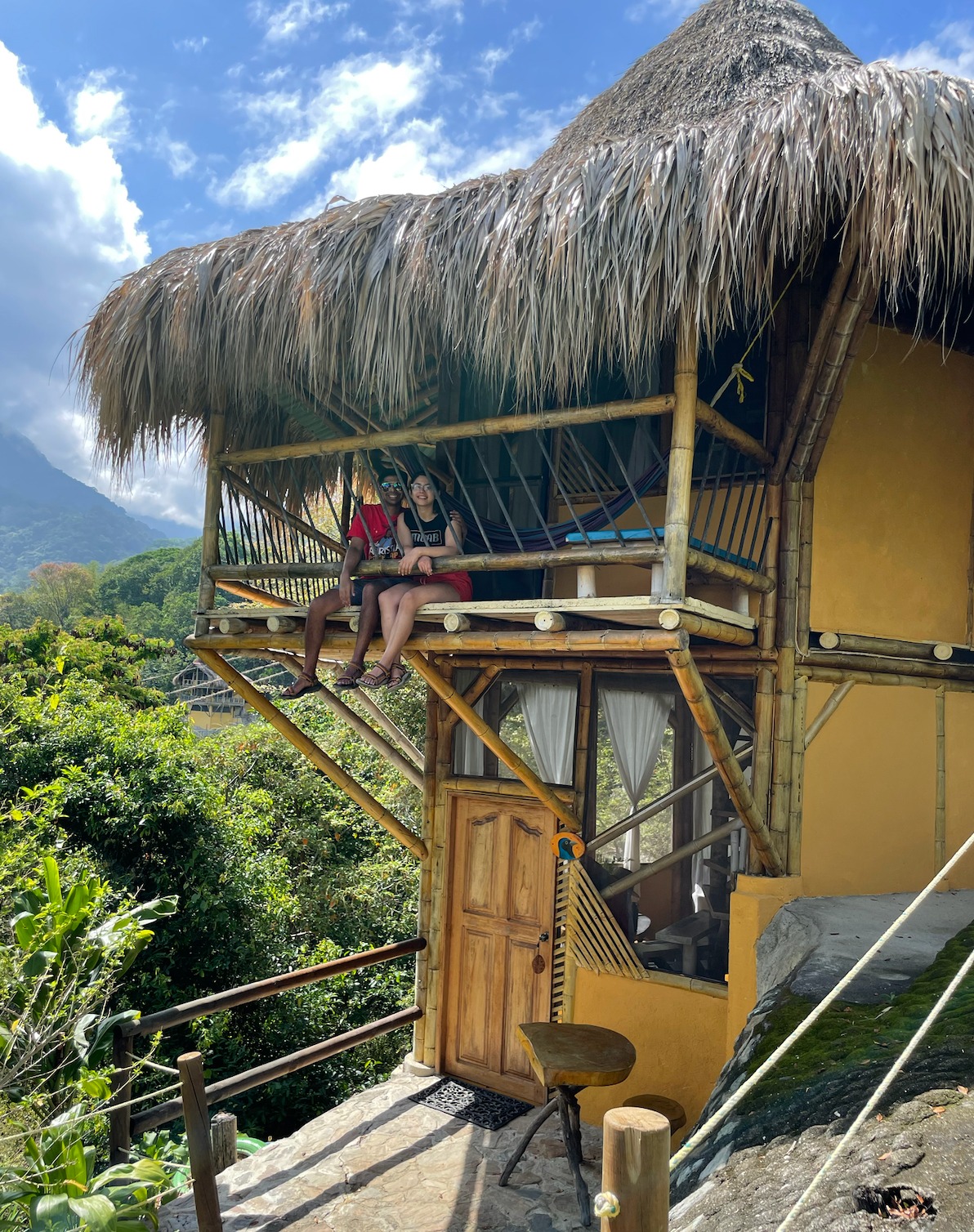 Two travelers sitting on the balcony of a thatched-roof bamboo treehouse above the Colombian jungle